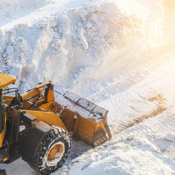 A large orange tractor removes snow from the road and clears the sidewalk. Cleaning and clearing roads in the city from snow in winter. Snow removal after snowfalls and blizzards.